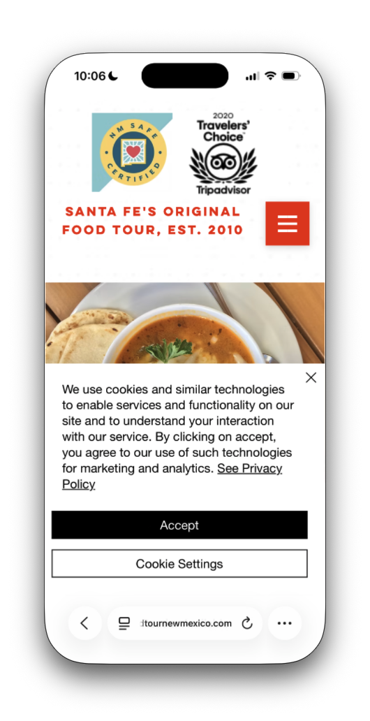 The Food Tour New Mexico site on an iPhone. At the top, an NM Safe Certified badge and a 2020 Tripadvisor Travelers’ Choice badge, then “Santa Fe’s Original Food Tour, est. 2010” and a menu button, then a photo of soup. A cookie notice with Accept and Cookie Settings buttons covers the bottom half of the screen.